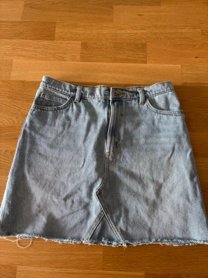 Ljusblå jeanskjol  - Snygg ljusblå jeanskjol från Monki med klassisk femficksdesign och rå, fransig kant nertill. Kjolen har dragkedja och knapp framtill samt bälteshällor. Perfekt för en avslappnad och trendig look.