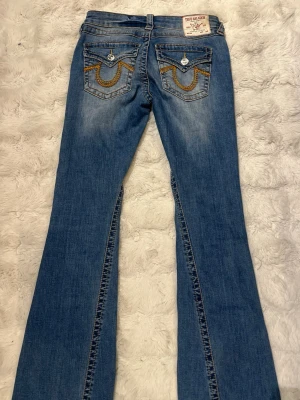 True Religion bootcut jeans blå - Snygga blå bootcut jeans från True Religion med ikoniska broderade fickor bak och slitningar framtill. Jeansen har klassisk femficksdesign, orange kontrastsömmar och är tillverkade i denim med lätt stretch. Perfekta för dig som gillar en avslappnad men trendig look.