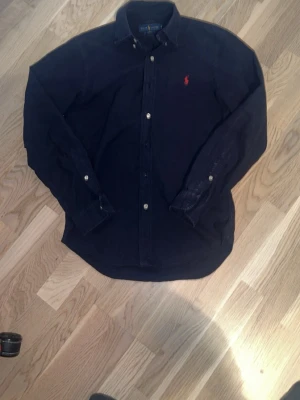 Mörkblå skjorta från Ralph Lauren - Klassisk mörkblå skjorta från Ralph Lauren med lång ärm och knappar framtill. Skjortan har en röd broderad logga på bröstet och är tillverkad i bomull. Perfekt för en stilren look.