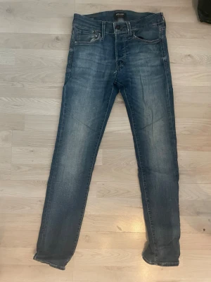 Blå slim fit jeans från Jack & Jones - Snygga blå slim fit jeans från Jack & Jones med klassisk femficksdesign  och slitningar för en cool, avslappnad look. Jeansen har en något tvättad finish och är tillverkade i bomull med lite stretch för extra komfort.   Jeansen är lite slitna och har två små hål därför säljer jag dom billigt. Hör av dig om du har några frågor! 🤩