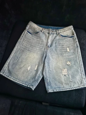 Ljusgrå denimshorts med slitningar - Säljer ett par ljusgrå denimshorts med råa slitningar och femficksmodell. Shortsen har en avslappnad passform och klassisk knappgylf. Perfekta för en chill och trendig look i sommar.