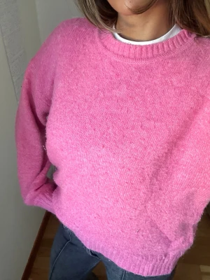 Rosa stickad tröja med rund hals - Mysig rosa stickad tröja med rund halsringning och ribbade muddar vid ärmslut och nederkant. Tröjan har en avslappnad passform och är perfekt att bära över en t-shirt eller skjorta. Lite nopprig som syns på bilden