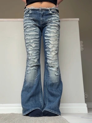 Jaded London bootcut jeans med slitningar - Säljer ett par blå bootcut jeans med kraftiga slitningar och blekta detaljer framtill. Jeansen har låg midja och klassisk femficksdesign. Perfekta för dig som gillar en edgy och trendig stil. Materialet är denim i bomull.