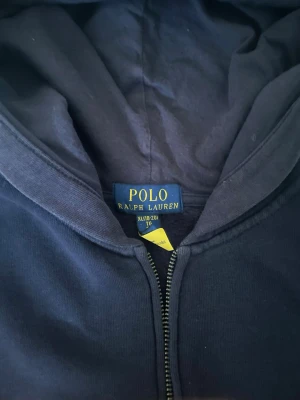 Mörkblå hoodie från Polo Ralph Lauren - Snygg mörkblå hoodie från Polo Ralph Lauren med dragkedja och klassisk röd broderad logga på bröstet. Hoodien har huva och är tillverkad i mjukt bomullsmaterial. Perfekt för en avslappnad och stilren look.