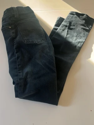 Mörkblå jeans från Nudie Jeans - Säljer ett par mörkblå jeans från Nudie Jeans i modellen 29/32. Klassisk femficksdesign med raka ben och snygga kontrastsömmar. Tillverkade i slitstark denim som passar perfekt till din vardagsstil. Scick 8/10 då knappt använda priset är ej hugget i sten och hör av er vid funderingar