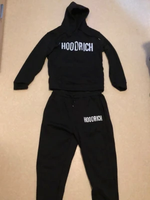 Hoodrich Tracksuit - Svart Hoodrich tracksuit bestående av hoodie med huva och matchande mjukisbyxor. Stilren branding som ger en tydlig streetwear-känsla 🔥  Hoodien har klassisk passform med huva och mjuk insida för komfort, medan byxorna har perfekt passform. Ett komplett set som passar perfekt för en clean och avslappnad streetstil 👌 