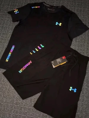 Svart träningsset från Under Armour - Snyggt svart träningsset från Under Armour med färgglada loggor och tryck. Setet består av en t-shirt och shorts, båda med regnbågsfärgade detaljer och UA-logga. Perfekt för gymmet eller sportaktiviteter. Materialet är lätt och andas.