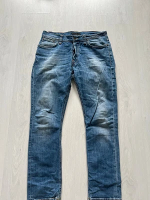 Blå jeans från Nudie Jeans - Säljer ett par klassiska blå jeans från Nudie Jeans med raka ben och snygga slitningar. Jeansen har fem fickor, orangea sömmar och en cool tvättad look. Perfekta för en avslappnad stil. Storlek 31/30