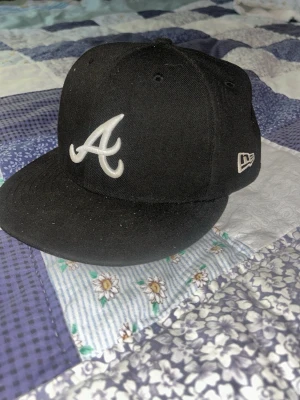 Svart New Era Atlanta Braves keps - Snygg svart keps från New Era med Atlanta Braves-logga broderad i vitt framtill. Klassisk flat peak och broderad MLB-logga baktill. Perfekt för dig som gillar streetwear och sportig stil.