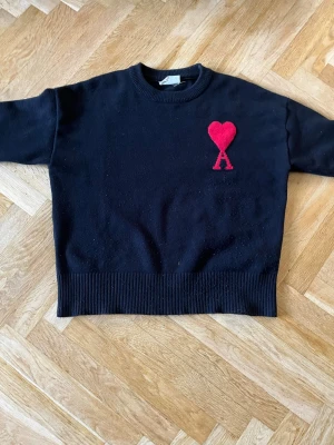 Svart stickad tröja från Ami Paris - PRIS KAN DISKUTERAS!   Svart stickad tröja från Ami Paris med röd hjärtlogga och bokstaven A broderad på bröstet. Tröjan har rund halsringning, ribbade muddar och en rak passform. Perfekt för dig som gillar stilrena plagg med en twist.