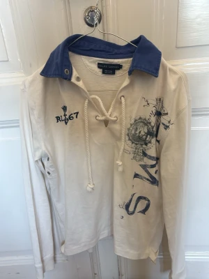 Vit långärmad skjorta från Ralph Lauren - Vit unik långärmad tröja från Ralph Lauren med blå krage, snörning i halsen och tryckdetaljer framtill. Skjortan har RL67-brodyr och marinblå grafiska inslag. Perfekt för en avslappnad och stilren look.