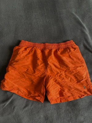 Orange bad shorts från Polo Ralph Lauren - Snygga orange bad shorts från Polo Ralph Lauren med klassisk broderad logga på benet. Modellen har elastisk midja och är tillverkad i ett lätt syntetmaterial, perfekt för varma dagar eller strandhäng.