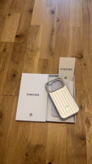 RIMOWA silverfärgat mobilskal iPhone Pro Max - Snyggt och stilrent mobilskal från RIMOWA i silver med grå kanter. Skalet har den klassiska RIMOWA-designen med räfflad yta och är tillverkat i tåligt plastmaterial. Perfekt för dig som vill ha ett exklusivt och modernt skydd till din mobil.