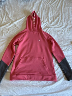 Rosa hoodie från Peak Performance - Cool rosa hoodie från Peak Performance med svart detalj på ärmarna och vit logga på bröstet. Huva med vit text längs kanten och stor magficka framtill. Perfekt för dig som vill ha en sportig och snygg look.