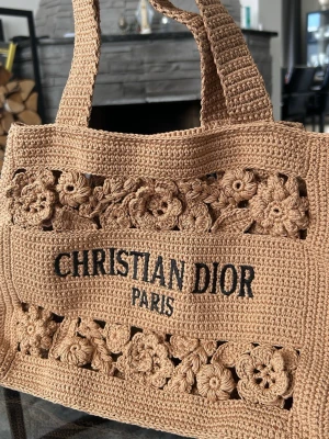 Beige virkad handväska  - Säljer en beige handväska, virkad i bomull för hand med blommiga detaljer och breda axelband. Väskan har ett unikt och trendigt utseende med tydlig logotyp framtill och snygga utskurna blommor. Perfekt som strandväska