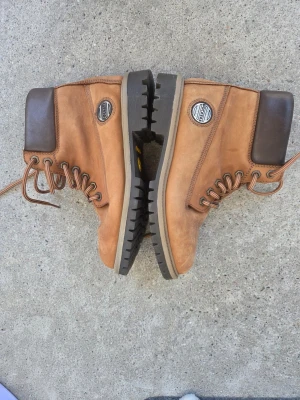 Bruna Dockers kängor i läder - Säljer ett par klassiska bruna kängor från Timberland med robust gummisula och mörkbrunt skaft. Kängorna är tillverkade i läder och har snörning framtill samt en rund Timberland-logga på sidan. Perfekta för dig som vill ha en tidlös och cool look.
