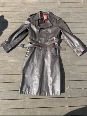 Lång brun skinnjacka med bälte - Säljer en lång brun skinnjacka med dubbelknäppning och bälte i midjan. Jackan har bred krage, axelklaffar och två snedställda fickor framtill. Perfekt för dig som vill ha en cool och tidlös look. Ena fickan behöver sys 