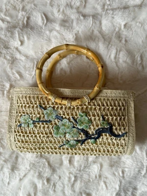 Stråväska med bambuhandtag och broderi - Snygg handväska i naturfärgat stråmaterial med runda bambuhandtag. Väskan har ett broderat blommönster i grönt och blått på framsidan som ger en unik och trendig look. Perfekt accessoar för dig som gillar boho-stil och naturmaterial.