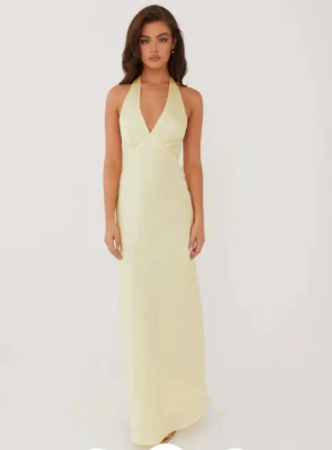 Ljus långklänning med halterneck - Elegant ljus gul långklänning med halterneck och djup v-ringning framtill. Klänningen har öppen rygg och knytband i nacken och baktill för en snygg siluett. Tillverkad i ett mjukt, glansigt material som ger en lyxig känsla.