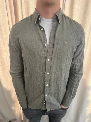 Grön linneskjorta från Morris - Snygg grön skjorta i linne med klassisk krage och vita knappar hela vägen. Skjortan har lång ärm och en liten broderad detalj på bröstet. Perfekt för dig som gillar en avslappnad men stilren look.