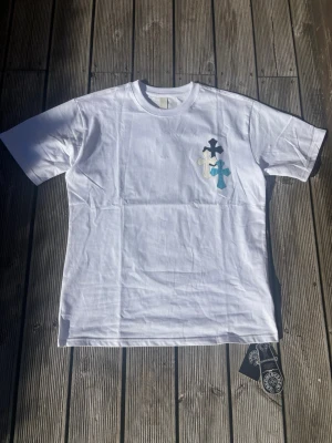 Chrome Hearts T-shirt - Säljer en vit t-shirt med flera korsapplikationer i svart, blått och silver på bröstet och ryggen. T-shirten har klassisk passform och korta ärmar. Perfekt för dig som gillar unika detaljer och streetwear-vibe.
