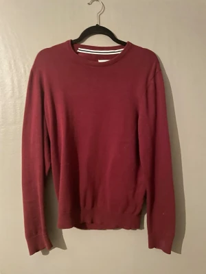 Mörkröd stickad tröja med rund hals - En enkel och stilren mörkröd stickad tröja med rund hals och långa ärmar. Tröjan har ribbstickade muddar vid ärmslut och nederkant. Perfekt för lager-på-lager och passar till jeans eller chinos. Materialet ser ut att vara mjukt och tunt.