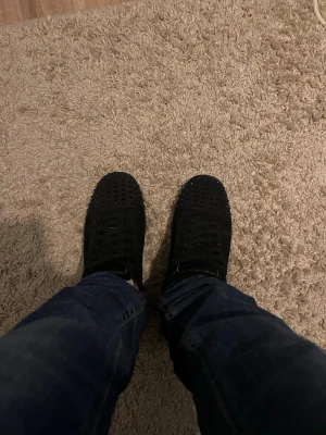 Svarta sneakers med nitar från Louboutin - Säljer ett par svarta sneakers från Christian Louboutin med coola nitar på tån och klassisk röd sula med logotyp. Skorna har snörning och är tillverkade i mocka för en lyxig känsla. Perfekt för dig som vill sticka ut med din stil. Kan sänka priset vid snabb affär 