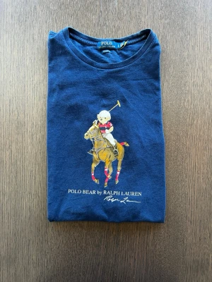 Blå Polo Bear t-shirt från Ralph Lauren - Snygg blå t-shirt från Ralph Lauren med Polo Bear-motiv på bröstet. Klassisk rund hals och korta ärmar. T-shirten är tillverkad i mjuk bomull och har en cool print med en björn som spelar polo. Perfekt för dig som gillar stilrena och ikoniska plagg. Storlek L men passar M