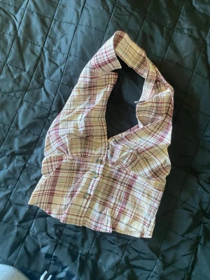 Rutigt halterneck linne med knappar - Säljer ett trendigt halterneck linne med röd, beige och brun rutigt mönster. Linnet har knappar framtill och en snygg passform som framhäver axlarna. Perfekt för dig som gillar retrostil och vill sticka ut med ett unikt plagg.