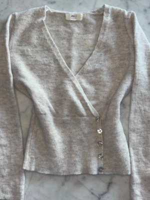 Beige omlottkofta från Gina Tricot - Säljer en beige omlottkofta från Gina Tricot i mjuk alpaca blend. Koftan har långa ärmar, djup v-ringning och dekorativa knappar på sidan. Perfekt för lager-på-lager och snygg att styla med jeans eller kjol.