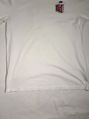 Vit Nike t-shirt med grafiskt tryck - Vit t-shirt från Nike med ett färgglatt grafiskt tryck på bröstet som visar en tecknad figur med sneakers, palmer och Nike-keps. Klassisk passform och korta ärmar, perfekt för dig som gillar streetwear och sneakers.