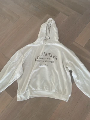 Vit hoodie med Los Angeles-tryck - Säljer en vit hoodie med tryck i brunt som lyder 'Los Angeles Downtown Sunset Boulevard CA, USA'. Hoodien har huva med dragsko och ribbade muddar. Perfekt för dig som gillar streetstyle och enkel design.