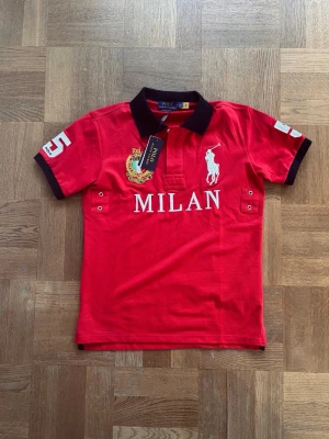 Röd Polo Ralph Lauren Milan skjorta - Cool röd kortärmad skjorta från Polo Ralph Lauren med svart krage och detaljer. Stort vitt Milan-tryck på framsidan, broderad logga och nummer 5 på ryggen. Perfekt för dig som gillar sportig stil och vill sticka ut.