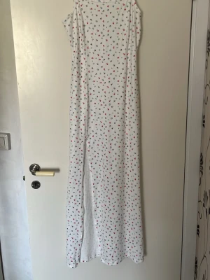 Långt vitt nattlinne med blommönster - Ett vitt nattlinne med smala axelband och ett långt, rakt fall. Tyg med små blommor i rött, blått och lila samt gröna blad. Perfekt för mysiga kvällar hemma. Materialet ser ut att vara mjukt och bekvämt, troligen bomull.