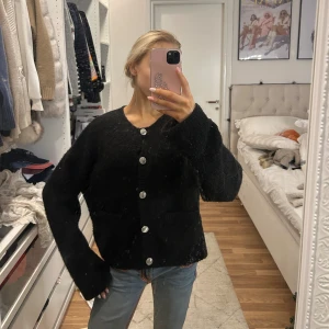 Svart kofta med stora knappar - Mysig svart kofta med stora silvriga knappar framtill och två fickor. Koftan har en rak och lite oversized passform med långa ärmar och rund hals. Från ginas premium kollektion