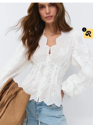 Vit broderad blus med spetsdetaljer - Superfin vit blus med broderade blommor och spetskantad v-ringning. Blusen har långärmade puffärmar och knappar framtill. Materialet är lätt och luftigt, perfekt för att skapa en romantisk look. Passar snyggt till jeans eller kjol.