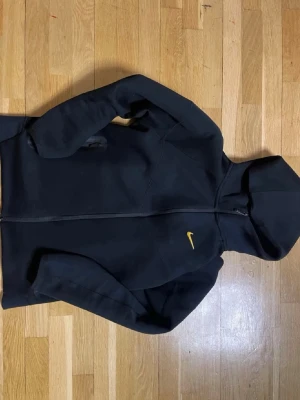 Svart Nike hoodie med gul logga - Svart hoodie från Nike med dragkedja och huva. Klassisk design med gul Nike-logga på bröstet. Tillverkad i mjukt material, perfekt för en avslappnad och sportig stil. Fickor på sidorna och bekväm passform. Står M i tröjan men är långt ifrån det i storleken, använder själv S och den är lite kort i armarna