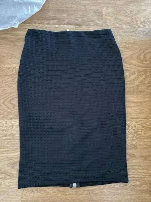 Svart pennkjol med dragkedja - Snygg svart pennkjol i strukturerat tyg med en lång silverfärgad dragkedja baktill. Kjolen har en tight passform och är knälång, perfekt till både vardags och mer uppklädda tillfällen. Materialet är stretchigt och ger en bekväm känsla.