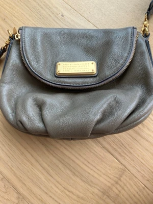Grå axelväska från Marc by Marc Jacobs - Jag säljer min snygga grå axelväska i mjukt läder från Marc by Marc Jacobs. Väskan har ett justerbart axelband, guldfärgade metalldetaljer och en dekorativ metallbricka på framsidan. Perfekt storlek för det viktigaste och en klassisk, rundad form. Väskan är väldigt väl omhändertagen och utan egentliga tecken på användning. Den är köpt direkt från butiken i USA. 