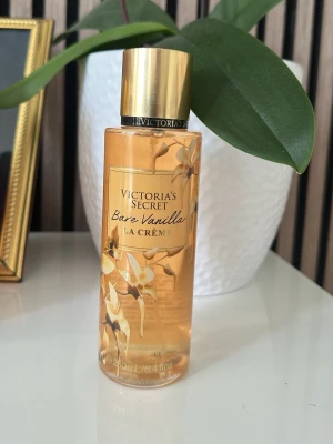 Victoria's Secret Bare Vanilla La Crème - En parfym från Victoria's Secret med doften Bare Vanilla La Crème. Flaskan är cylindrisk och transparent med guldiga detaljer och blommönster. Guldigt skruvlock och elegant design. Perfekt för dig som gillar vaniljtoner och lyxig känsla.