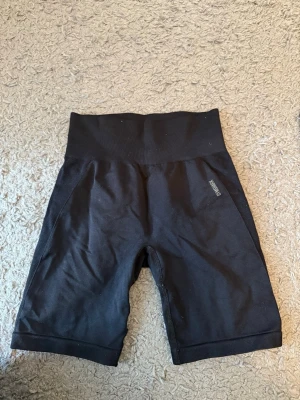 Svarta träningsshort från Gymshark - Säljer ett par svarta gym shorts från Gymshark med hög midja och logotyp på baksidan. Shortsen är tillverkade i ett stretchigt syntetmaterial och har en tight passform som är perfekt för gym och träning.