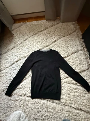 Svart tröja från j.lindeberg - Snygg svart tröja från Calvin Klein med rund halsringning och diskret logga på bröstet. Tröjan har lång ärm och är tillverkad i ett mjukt, tunt material som passar perfekt till lager på lager. Enkel och stilren design som funkar till det mesta.