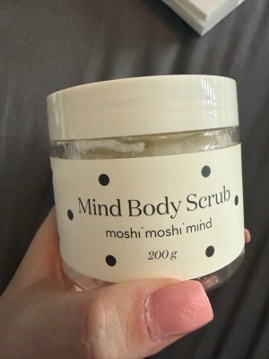Mind Body Scrub moshi moshi mind - Mind Body Scrub från moshi moshi mind i en genomskinlig burk med vit etikett och svarta prickar. Innehåller 200 g kroppsskrubb som exfolierar huden. Perfekt för dig som vill ha en fräsch och mjuk känsla efter duschen.