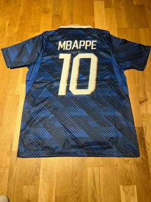 Frankrike Mbappé matchtröja blå - Säljer en blå matchtröja från Nike med Frankrikes landslagsemblem och nummer 10 Mbappé på ryggen. Tröjan har vita detaljer, klassisk krage och är tillverkad i lätt, ventilerande material. Perfekt för dig som älskar fotboll och vill stötta Frankrike.