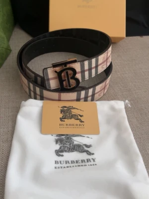 Burberry bälte med TB-spänne - Säljer ett exklusivt Burberry bälte med klassiskt rutigt mönster och ett elegant TB-spänne i metall. Bältet har beige, bruna och svarta toner och är tillverkat i skinn. Perfekt för att lyfta din outfit med en ikonisk accessoar.