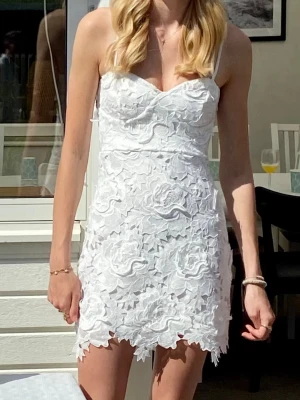 Vit spetsklänning med blommönster - Kort vit klänning med vackert blommigt spetsmönster och figurnära passform. Klänningen har smala axelband, omlottliknande byst och dekorativt avslut nertill. Perfekt för sommarens alla tillfällen.