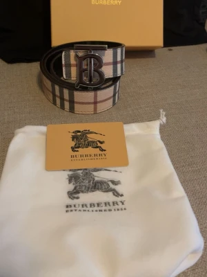 Rutmönstrat Burberry bälte i skinn - Snyggt Burberry bälte i skinn med klassiskt rutmönster i beige, svart och rött. Stort metallspänne med Burberrys logotyp och detaljerad gravyr på bältet. Perfekt accessoar för att lyfta din outfit.
