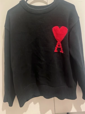 Svart stickad tröja Ami Paris - Svart stickad tröja från Ami Paris med röd hjärt- och A-detalj på bröstet. Tröjan har rund halsringning, ribbade muddar och avslut. Perfekt för dig som gillar stilrena plagg med en twist.
