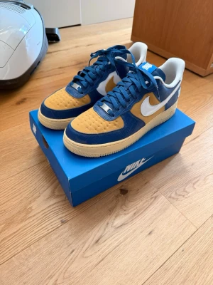 Nike Air Force 1 Low SP ”Undefeated 5 on it blue yellow” - Nike Air Force 1 x Undefeated sneakers med blå och gula paneler, vit swoosh och detaljer. Lågt skaft, klassisk siluett och robust gummisula. Materialmix av mocka och läder, med Undefeated-logga på hälen. Perfekt för dig som vill sticka ut med stil.