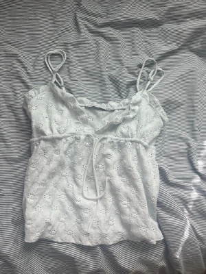 Vit spetslinnetopp från 157 - Söt vit linnetopp från 157 med broderad spets och tunna axelband. Toppen har dekorativ knytning framtill och ett romantiskt blommönster i tyget. Perfekt för sommarens varma dagar och ger en fräsch look.